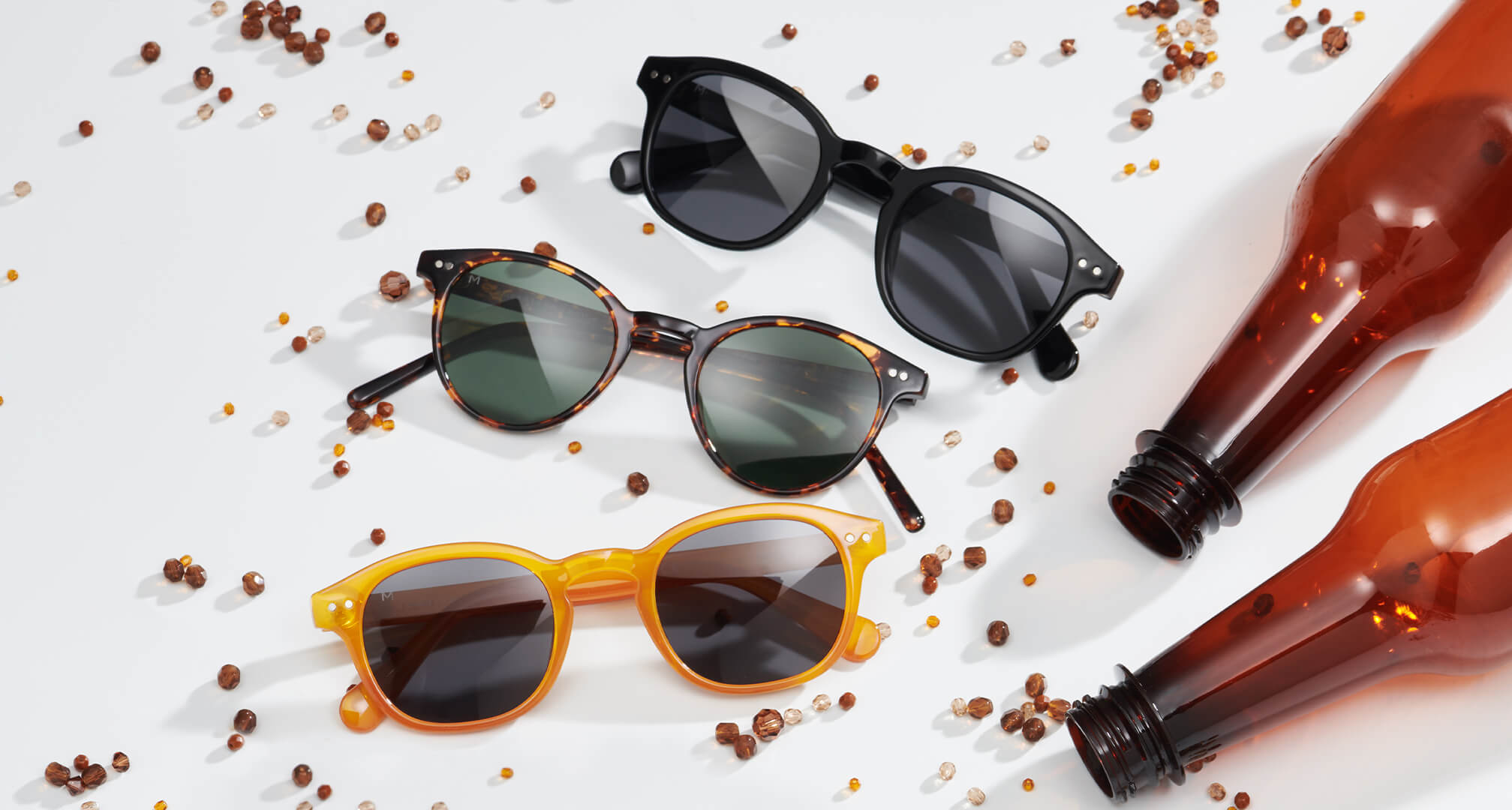 kolm paari jätkusuutlikest materjalidest valmistatud päikeseprille valgel laual, ümberringi pärlid ja plastpudelid.